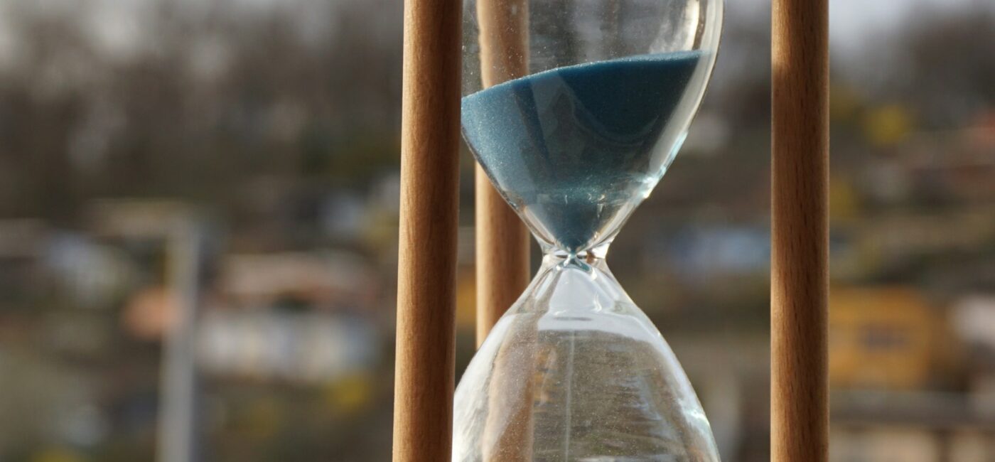 Zeitperspektiven im Coaching: Vergangenheit, Gegenwart und Zukunft Eine Sanduhr mit blauem Sand steht in einem Holzrahmen vor unscharfem Hintergrund.